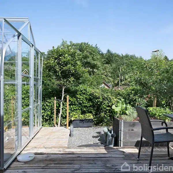 Et drivhus står stille på en træterrasse ved siden af et sæt havemøbler. I baggrunden ses en frodig have med træer og grønt løv under en klar blå himmel.