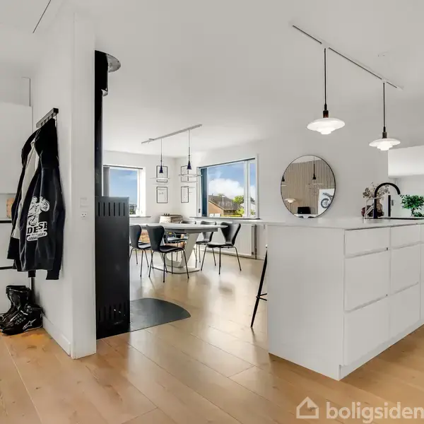 Entre med åbent køkken og spiseområde i moderne design; lyse trægulve og hvide vægge. Runde lamper hænger over køkkenøen, og der er udsigt gennem store vinduer. En frakke og støvler er ved indgangen.