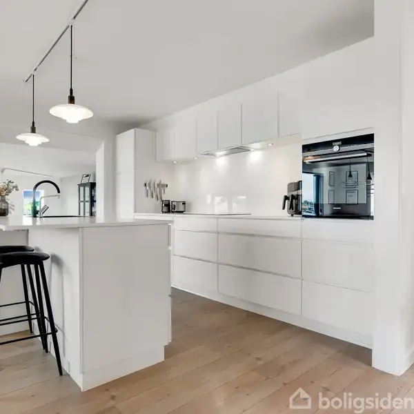 Et moderne køkken med hvide skabe og en indbygget ovn til højre. En køkkenø med to sorte barstole er midtpunkt. Rummet er lyst og åbent.