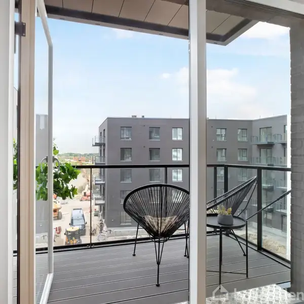 En balkon med to sorte stole og et lille bord står i en moderne lejlighedsbygning. Udsigten viser flere grå bygninger og en klar blå himmel.
