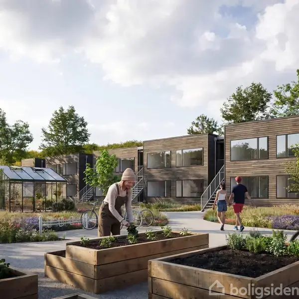 En person arbejder i en hævet plantekasse i en moderne boligkompleks gårdhave med cyklister og kontormoduler i baggrunden. Området er omgivet af grønne træer og planter.