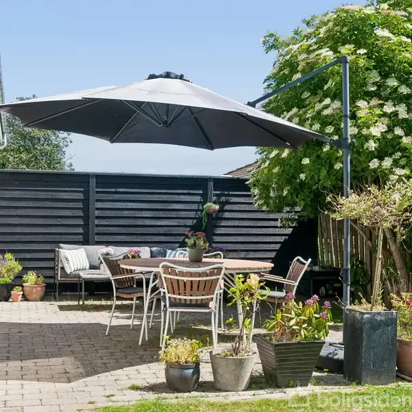 Terrasse med havebord og stole under en stor parasol. Omgivet af blomster i potter og grønne planter. Sort hegn til højre og en hvid husvæg til venstre.