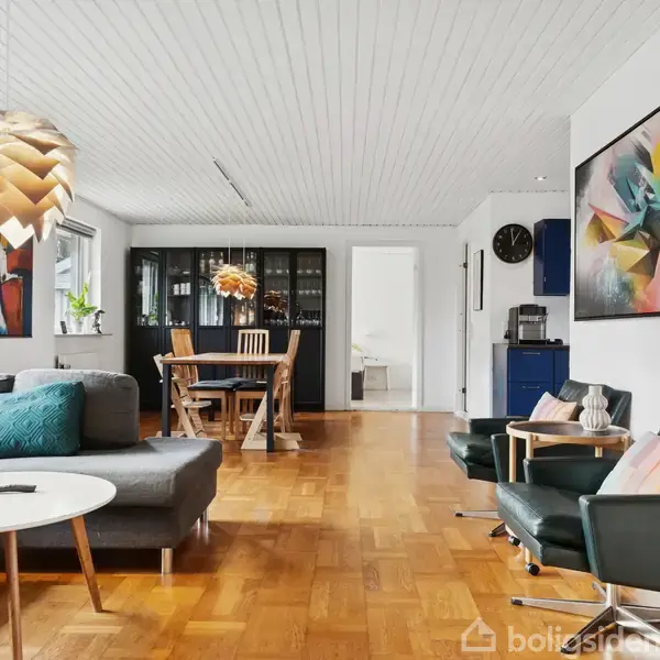 Sofa står i stue med trægulv og kunstværk på væggen. Et spisebord ses i baggrunden under en moderne loftlampe. Rummet har store vinduer og elegant indretning.