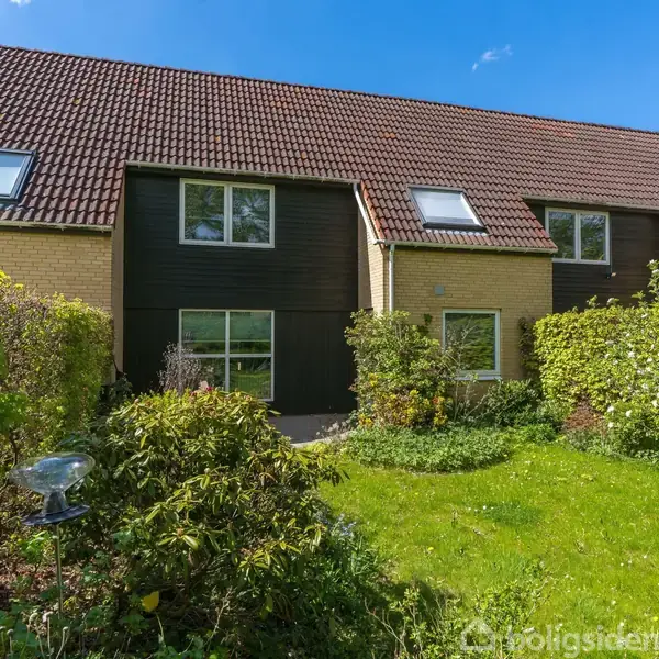 Rækkehus med rødt tag observeres i en frodig have med grøn plæne og buske omkring. Sollys skinner på bygningens gule mursten og sorte facade.