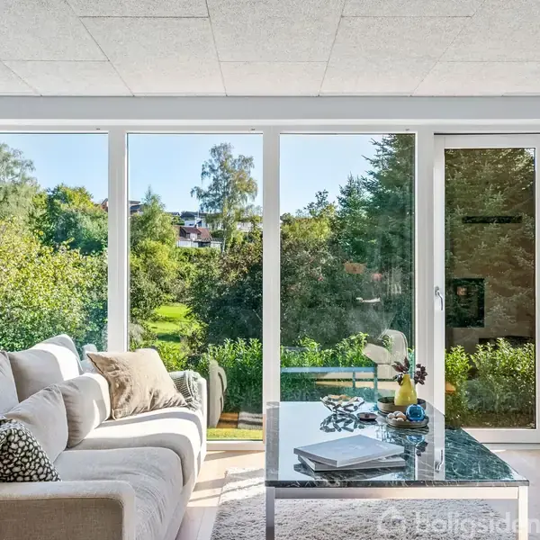 Sofa med puder står foran store vinduer, der giver udsigt til frodig have. Et sofabord med bøger og dekorationer står i centrum. Fjernsynet hænger på højre væg.