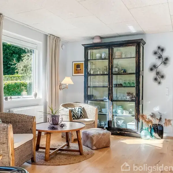 En glasvitrine fyldt med glas og service står i et lyst stuehjørne med træmøbler og bløde tekstiler. Et stort vindue lukker naturligt lys ind, omgivet af grøn beplantning udendørs.
