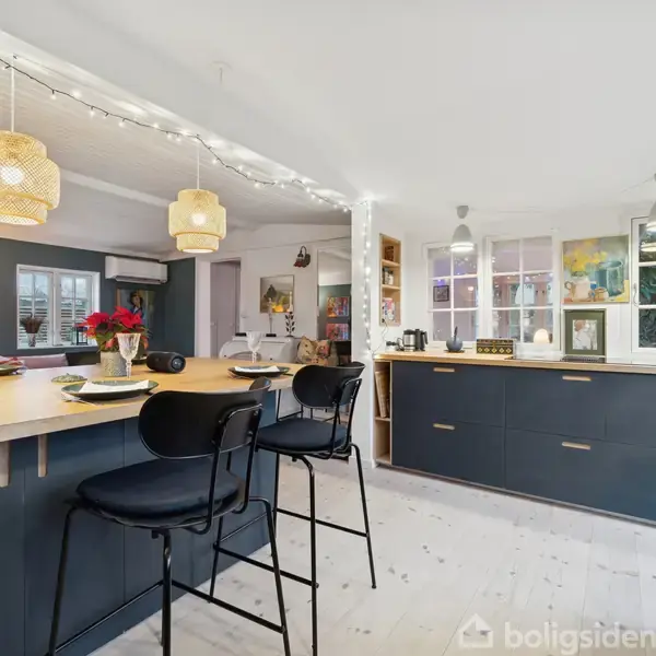 Et stort køkken med sorte barstole ved en lys køkken-ø; lamper hænger over bordpladen. Vinduet og skabe i baggrunden, dekoreret med billeder og lys.