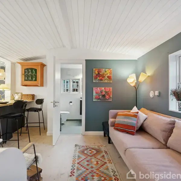 Stue med en beige sofa fyldt med farverige puder ved et vindue. Midt i rummet er en tæppebelagt gulv og vægkunst. Til venstre ses et åbent køkken med barstole.