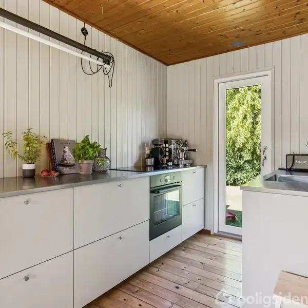 Et køkken med grå skabe og træloft. En ovn er integreret i skabene, og en vask er placeret ved et vindue, der åbner mod en grøn have. Brætgulv og hvide vægge.