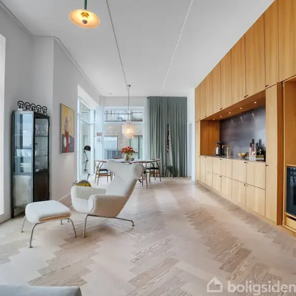 En moderne lejlighed med åbent rum; en lænestol står ved et stort vindue. Indbyggede skabe og ovn er til højre. Spisebord ses i baggrunden med blomster.