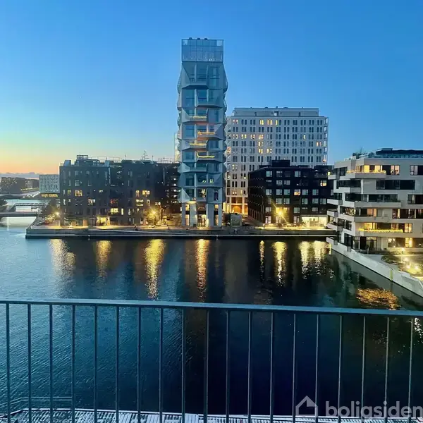 Bygninger reflekterer i vandet, mens skumringen skaber en rolig atmosfære i en moderne bydel med et tårnhøjt, unikt designet byggeri i midten omgivet af andre lejlighedskomplekser.