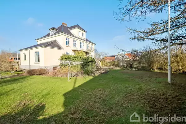 En stor, lys villa står stille i en frodig have, omgivet af træer og buske. Flagstangen står til højre på den grønne græsplæne under en klar, blå himmel.