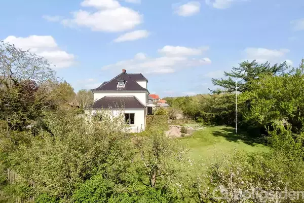 Et to-etagers hus med sort tag ligger midt i en frodig have. Omkring ejendommen ses en grøn græsplæne og høje træer, under en klar blå himmel med skyer.