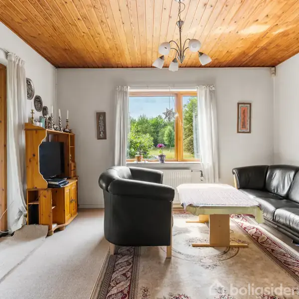 Stue med sort lædersofa og lænestol omkring et bord. Vindue med udsigt til have. Træloft og lyst tæppe oplyst af en lysekrone. Møbler inkluderer et fjernsyn på reol.