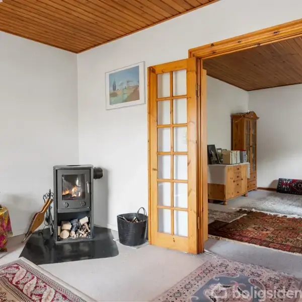 En fritstående brændeovn står i et hyggeligt rum med tæpper på gulvet. Rummet grænser op til en anden stue gennem en glasdøråbning. Møbler og dekorationer skaber en hjemlig atmosfære.