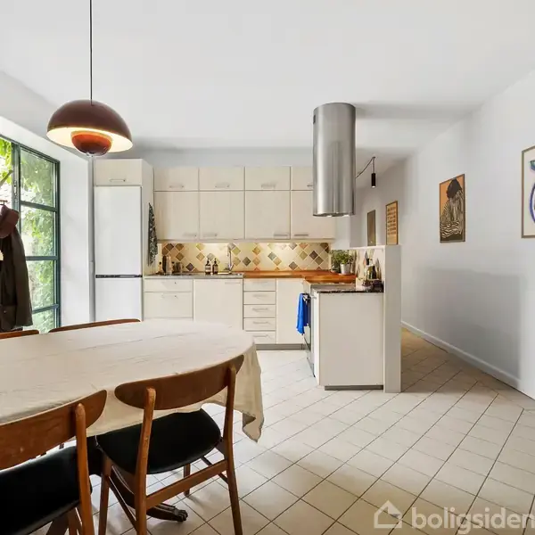 Spisebord med dækket hvid dug og buket blomster står i køkken. Køkkenet har lyse skabe, kogeplade, ovn og emhætte. Væggene prydes med billeder.