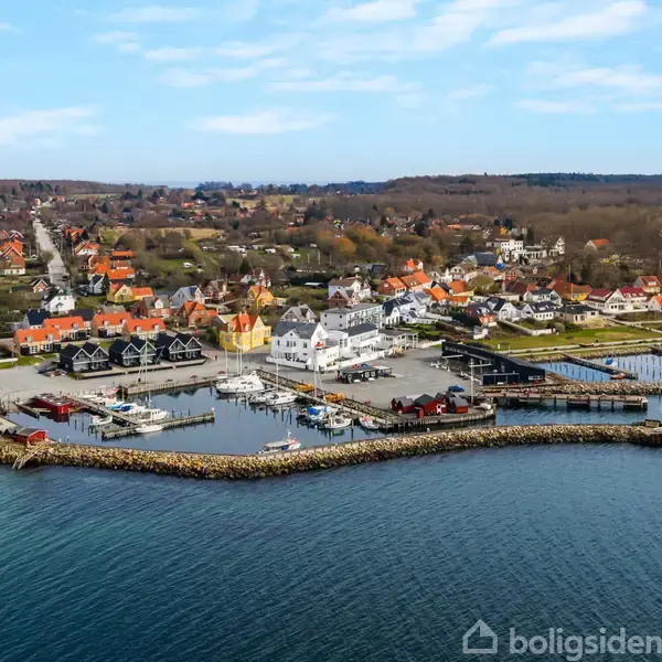 Havn med både ligger stille i vandet omgivet af en kystby med mange rødtagehuse og grønne områder. Vejr er klart med blå himmel og spredte skyer.