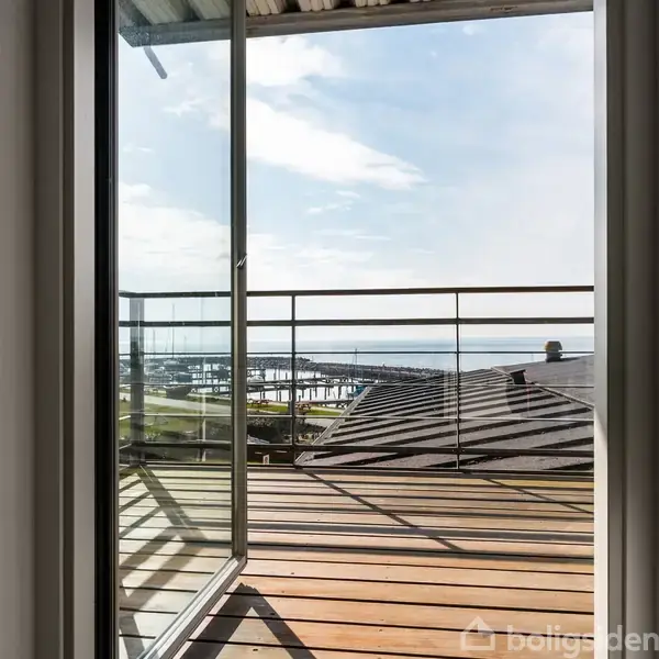 En åben altandør leder ud til en træterrasse med udsigt over havnen og havet, mens en blå himmel med spredte skyer giver lys og luft.