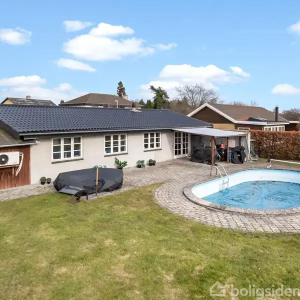 En bungalow med sort tag ligger i en have med en oval swimmingpool og flisebelagt terrasse. Omkranset af hække og nabohuse under en blå himmel med spredte skyer.
