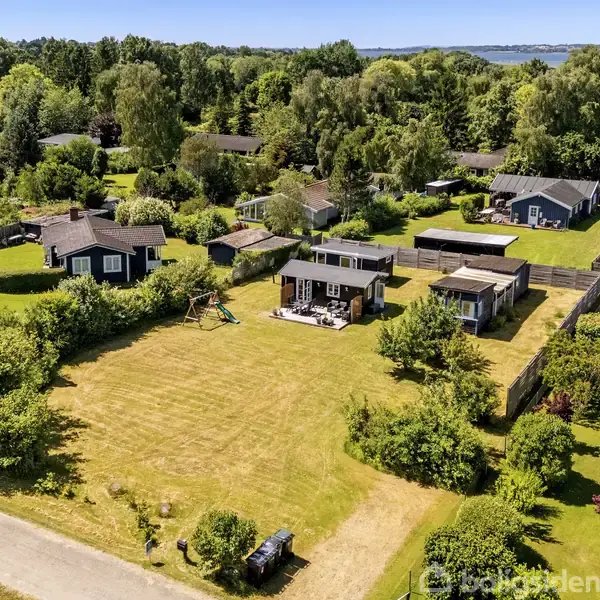 Et stort sommerhus med en græsplæne ligger mellem andre huse i et frodigt grønt område. Der er skov omkranset landskab og et glimt af vand i baggrunden.