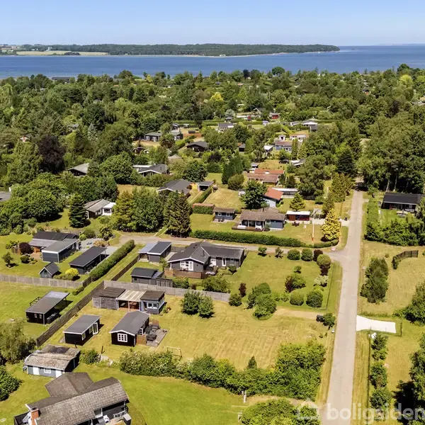 Huse står spredt i en grøn, skovklædt landsby nær en vej. I baggrunden ses en bred fjord eller sø.
