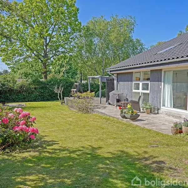 Et hus står i en frodig have med grønne træer og buske omkring. Der er en terrasse med havemøbler foran huset, og blomsterbed pryder græsplænen.