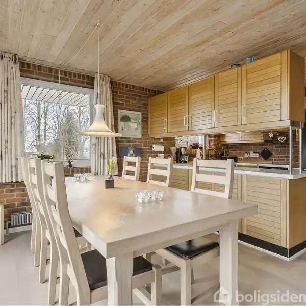 Langt spisebord med seks stole i en spisestue. Bordet er dækket med nogle lys og en vase med blomster. Rummet har murstensvægge, træloft og et stort vindue med gardiner.