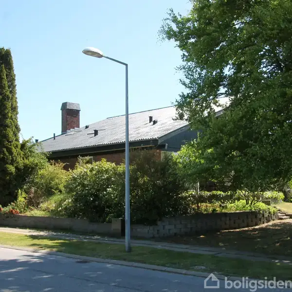 En villa med skråt tag omgivet af grønne træer og buske, placeret ved en vej med en gadelampe og en parkeret bil til venstre.