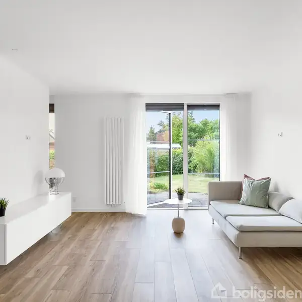 Stue med en sofa placeret langs højre væg, vendt mod en stor glasdør med udsigt til en grøn have. Trægulv og en hvid skillevæg til venstre med indbygget hylde.