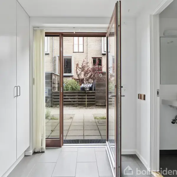 En glasdør åbner ud til en flisebelagt terrasse med planter i en rækkehusgård. Indenfor ses indbyggede skabe til venstre, og et lille badeværelse med vask til højre.