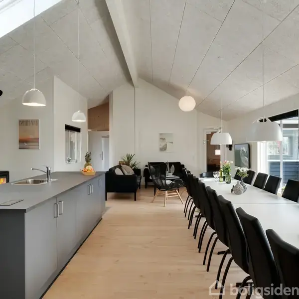Køkken-alrum med sort køkkenbord, siddepladser til mange ved hvidt langt spisebord. Lyse vægge og træloft, moderne lamper og vinduer, lysindfald fra ovenlysvindue.