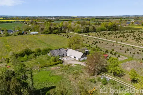 En gård ligger stille på en grøn mark med flere bygninger omkring, omgivet af træer og marker, der strækker sig til horisonten, under en klar, blå himmel.