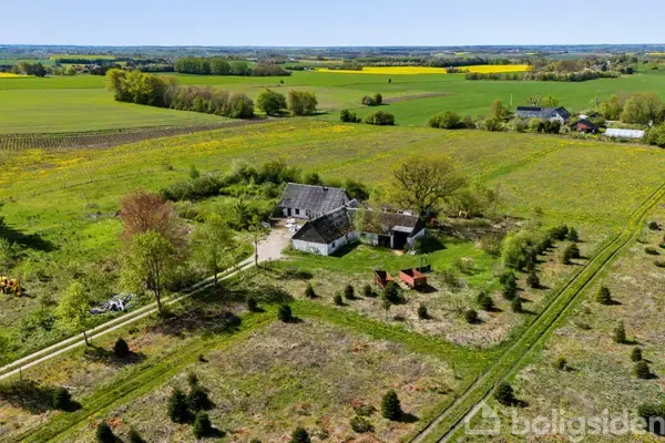 Gårdkompleks med flere bygninger placeret på en stor grund. Bygningerne omgivet af marker og få træer. Landejendom beliggende i et åbent, landligt område med udsigt til horisonten.