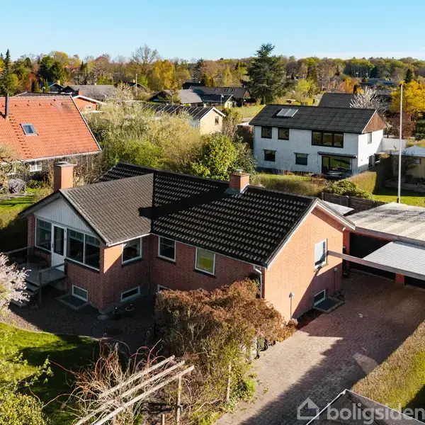 En rødstensvilla med sort tag står i en grøn, frodig have. Ved siden af huset er en dobbelt carport. Omkringliggende huse ses i en solrig beboelseskvarter.