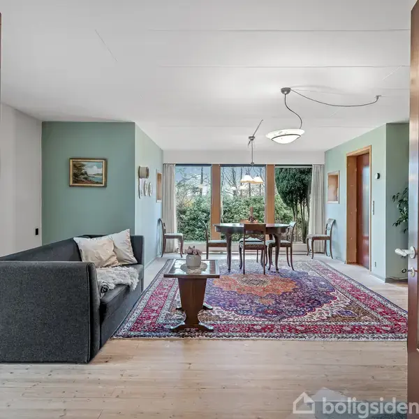 Gråt sofaområde åbner til en spisestue med et træbord og stole. Store vinduer slipper lys ind, og farverige tæpper dækker trægulvet i et grønt indrettet rum.