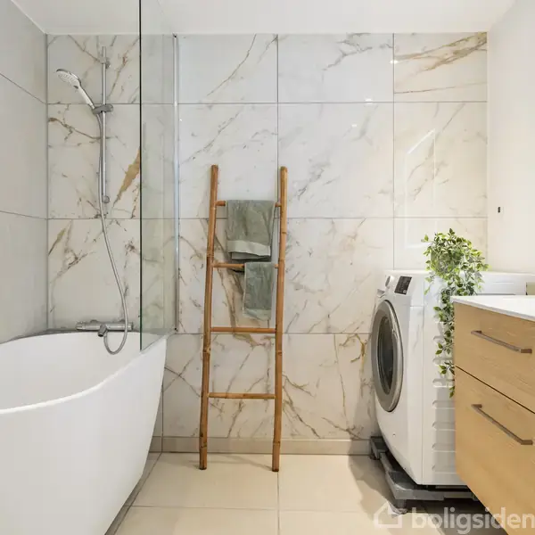 Badekar står mod en marmoreret væg ved siden af et håndklæde på en stige. En vaskemaskine er placeret ved træskuffemøbler med en plante og spejl over.