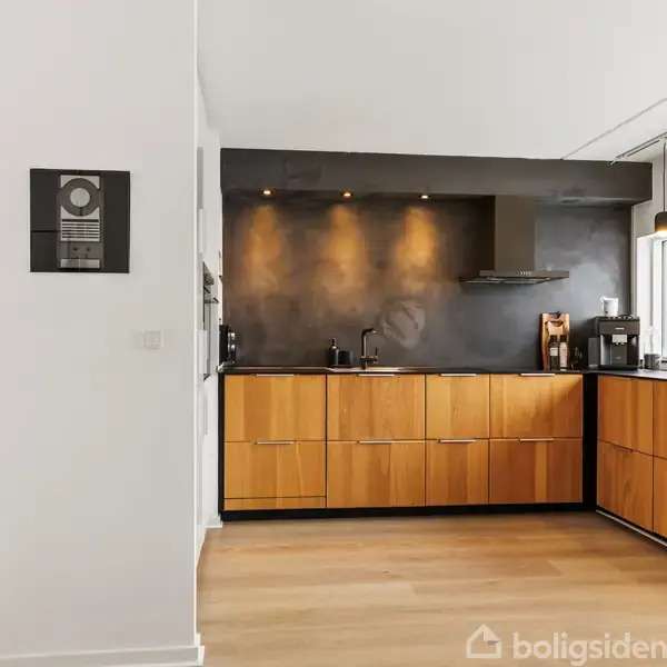 Køkken med træskabslåger og mørk væg. Overfladerne er minimalistiske med få køkkenapparater. Rummet har en stor rude, der lukker lys ind og pendellamper hænger fra loftet.