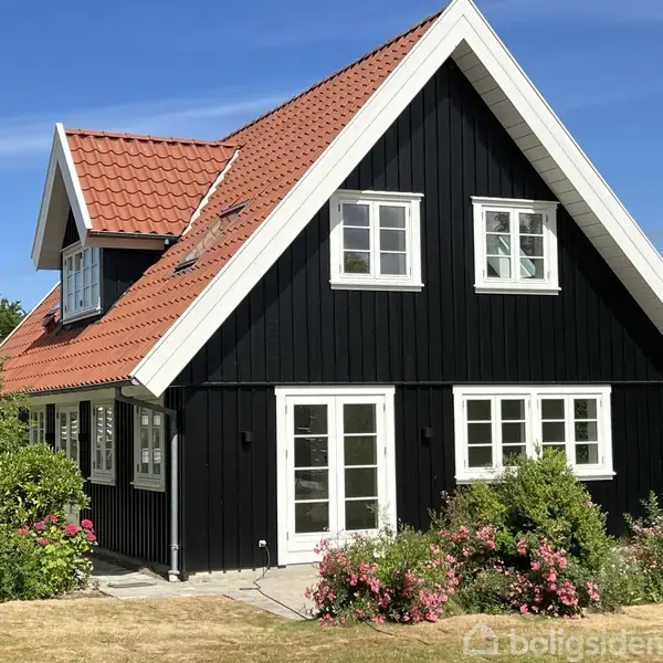 Et sortmalet træhus med rødt tegltag står omgivet af grønne buske og blomster. Solen skinner, og der er blå himmel i baggrunden.