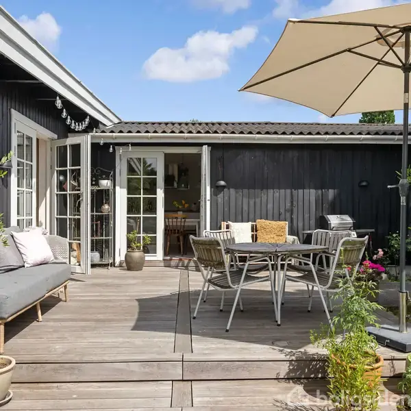 Terrasse med træmøbler arrangementet omkring et spisebord under en stor parasol. En sofa med puder står ved siden af. I baggrunden ses et sortmalet hus med åbne franske døre.