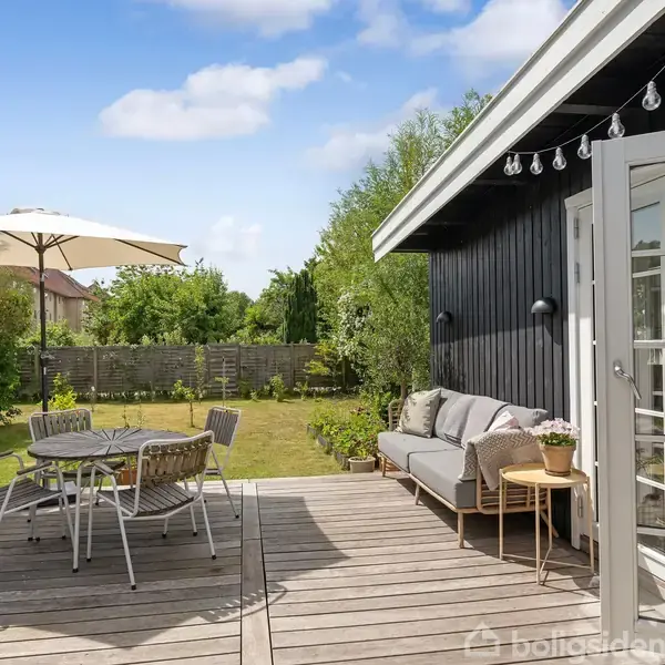 Terrasse med havemøbler og parasol. Sofa står ved siden af glasdør. Træterrassen ligger ved huset og grænser op til en græsplæne med hegnet have. Klar blå himmel.