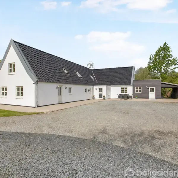 Hvidt, vinklet hus med sort tag står på grusplads omgivet af grønne træer og buske. Ingen tekst i billedet.