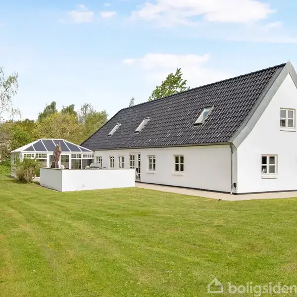 Hvidt hus med sort tag står roligt i en grøn have med græsplæne. I baggrunden findes et drivhus ved siden af huset, omgivet af træer under en klar blå himmel.