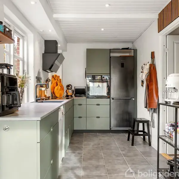 Køkken med grønne skabe, kaffemaskine og køleskab i baggrunden. En hylde med genstande hænger på væggen. Rummet har grå flisegulv og hvidt loft med indbyggede spots.