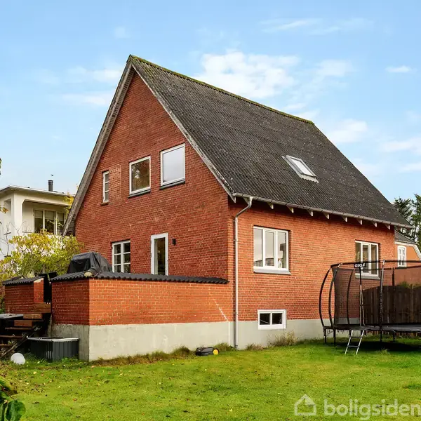 En rød murstensvilla med skråt tag står i en grøn have. En trampolin er placeret på græsset ved siden af huset. Der er nogle buske og træer i baggrunden.