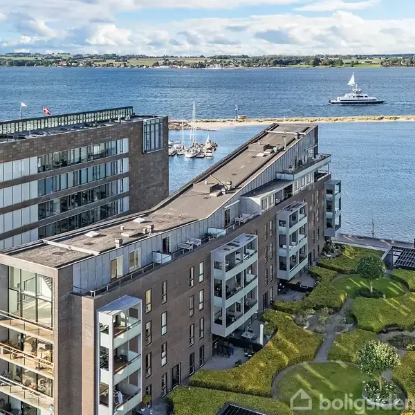 Moderne lejlighedskompleks med altaner og fladt tag, omgivet af grønne områder. I baggrunden ses en rolig havnefront med både og en mole, samt en sejlbåd på vandet.