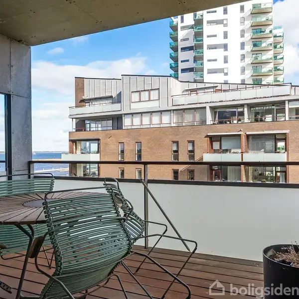 Et rundt havebord med metalstole står på en altan med glasrækværk. I baggrunden ses moderne lejlighedsbygninger og delvist udsigt til havet under en blå himmel.