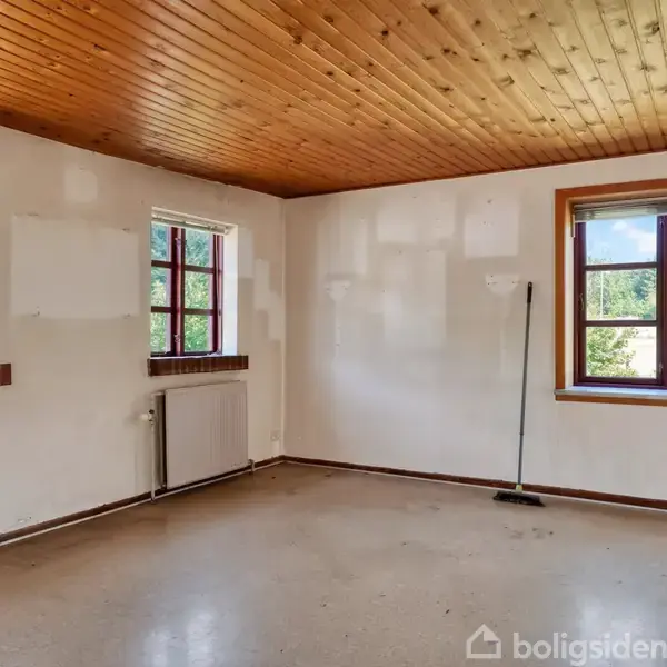 Et tomt rum med lyst trægulv; en kost står op ad væggen. Værelset har hvidmalede vægge, træloft og tre vinduer med udsigt til grønne omgivelser.