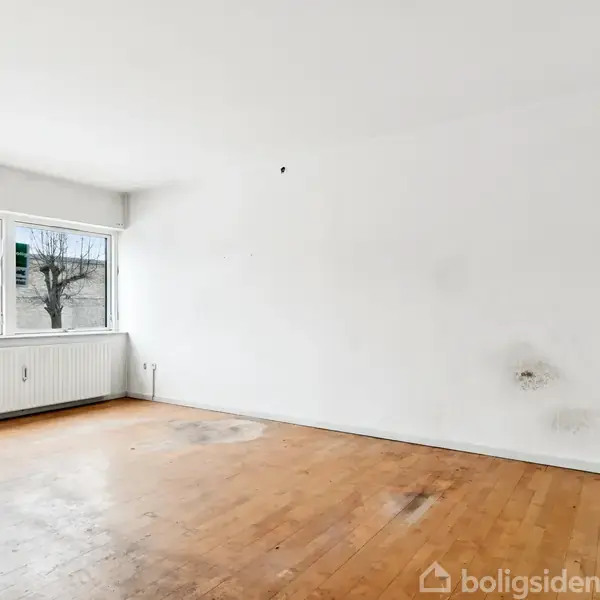 Et tomt værelse med et trægulv, hvide vægge og et stort vindue til venstre, udenfor ses træer uden blade. Radiator under vinduet. Ingen tekst i billedet.