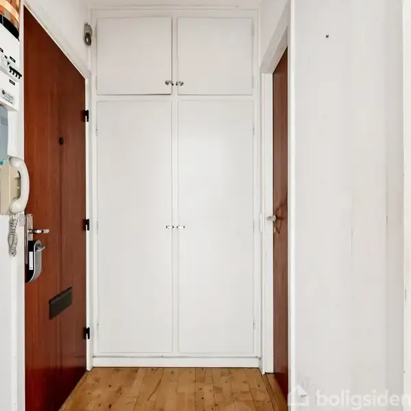 Hvid skabsvæg står stille i en snæver entré med trælaminatgulv. Til venstre er der en dørtelefon og elmålere, mens hoveddøren er brun med et postindkast.