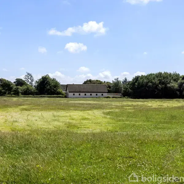 En hvid bygning står stille omgivet af en stor, grøn mark. Træer omkranser området, og himlen er let overskyet med hvide skyer. Ingen tekst er til stede.